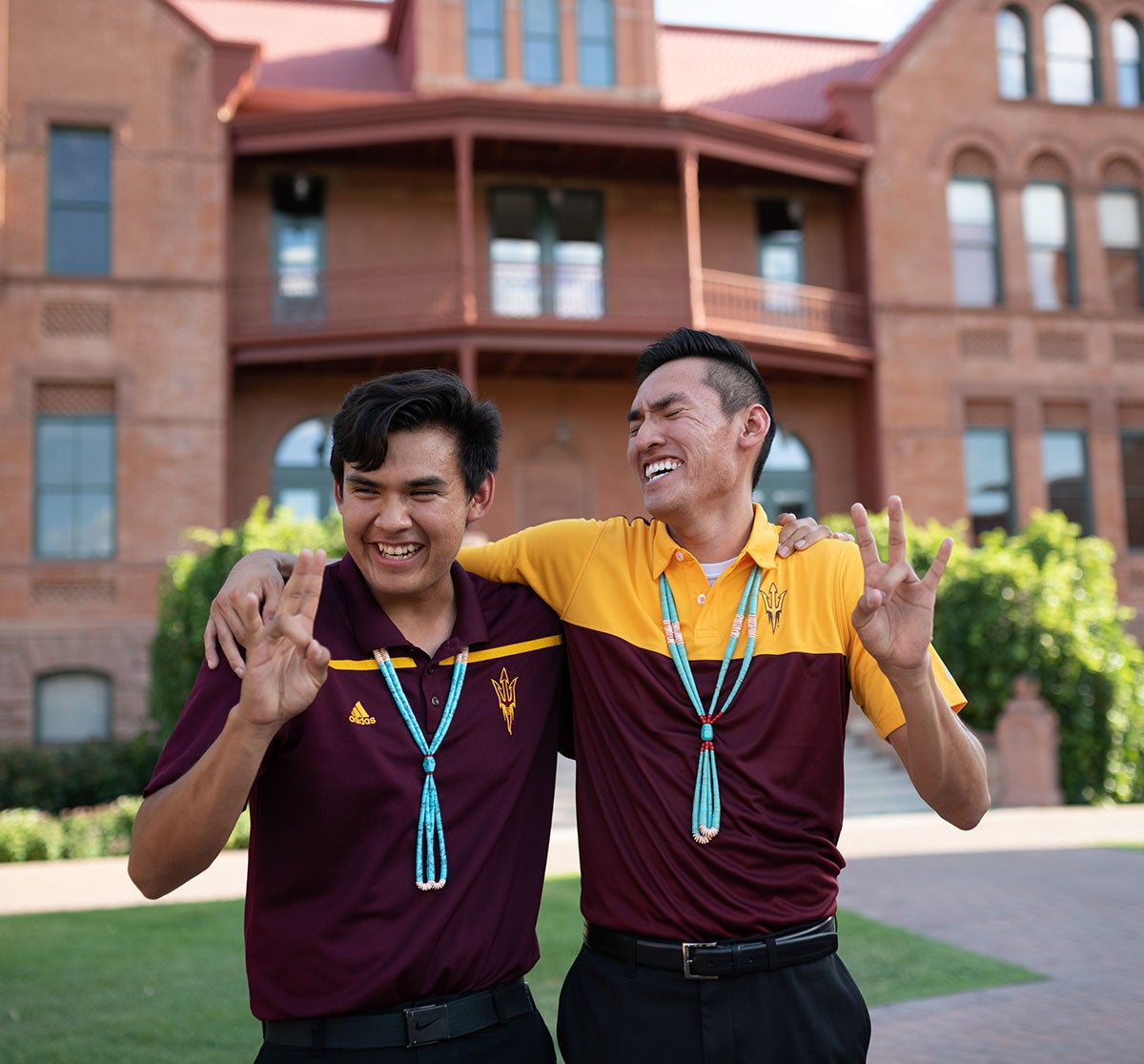 Two students near old main building forks up hand sign