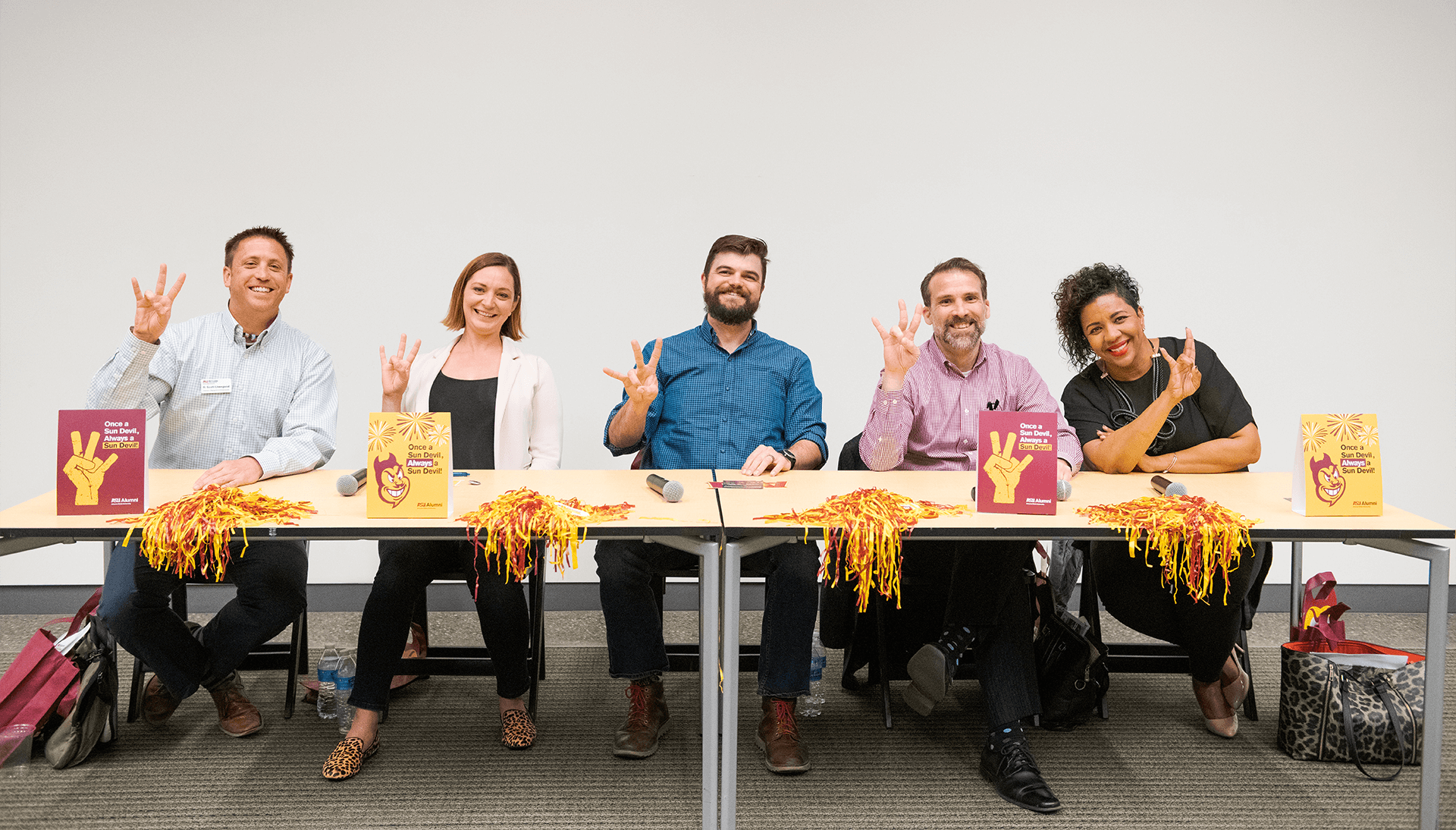 A photograph of five ASU alumni seated panel-style across a long table, holding their hands up with the ASU pitchfork symbol. Signs on the table read "Once a sun devil, always a sun devil."